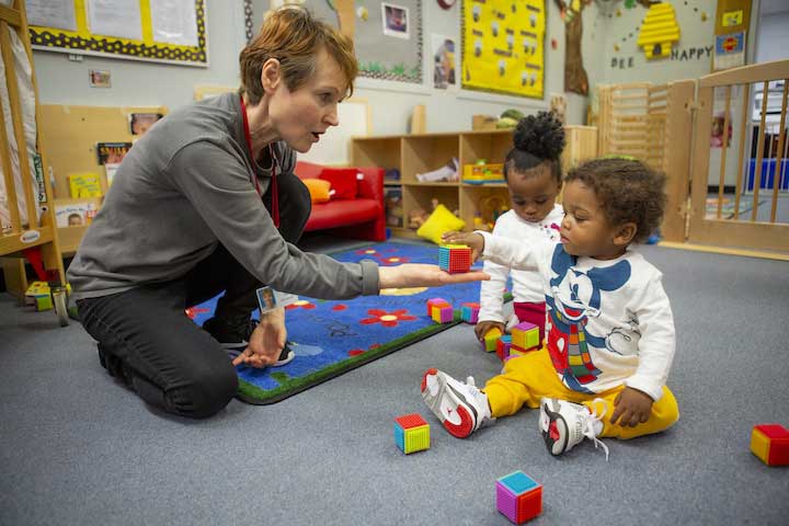 Teacher and toddlers play on carpet. Teacher holds a block in her hand as a child reaches for it